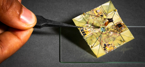 Small, square microflier device being held by tweezers over a piece of glass